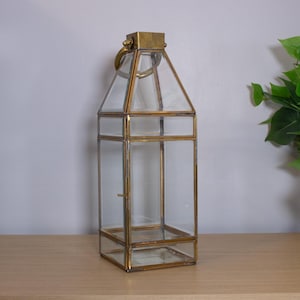 May include: A tall, rectangular lantern featuring a brass metal frame and clear glass panels. The lantern has a tapered top section, a hinged side door with a small latch, and a circular brass ring handle attached to the top cap.