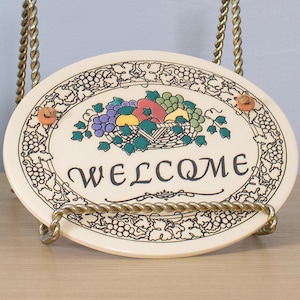 May include: Oval ceramic welcome sign with a fruit basket design in the center. The sign has a cream-colored background with a border of grapes. The word "WELCOME" is written in black script.