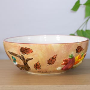 May include: A ceramic bowl with a cream-colored interior and a tan exterior. The bowl is decorated with hand-painted autumn leaves, a bird, and vegetables. The word "Elaine" is written on the bowl.