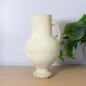 May include: A cream-colored ceramic pitcher with a rounded body, a narrow neck, and a single curved handle. The vessel features a textured, matte surface with subtle horizontal ridges and a flared base, standing upright with a simple, functional design.