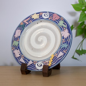 May include: A decorative ceramic plate with a blue border featuring colorful abstract shapes. The plate has a white and gray spiral design in the center and is displayed on a wooden stand.