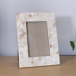 May include: A rectangular picture frame featuring a decorative border composed of small, iridescent white and cream-colored mother-of-pearl tiles arranged in a woven, crisscross pattern. The frame has a central rectangular opening for a photo.
