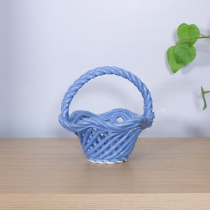 May include: A light blue ceramic basket with a woven design and a braided handle. The basket sits on a wooden surface, with a white wall and green plant in the background. The basket is approximately 6 inches tall.