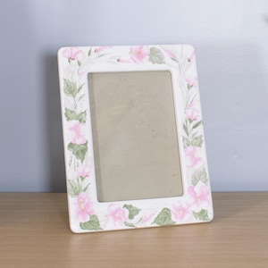 May include: A rectangular ceramic picture frame with a white base featuring a hand-painted floral pattern of pink hibiscus flowers and green leaves surrounding the central opening. The frame has a smooth, glossy finish and a vertical orientation.