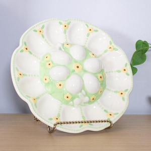 May include: A white ceramic egg plate with a scalloped edge, decorated with yellow flowers and a green center. The center holds ceramic eggs. The plate is displayed on a gold-colored metal stand. A decorative piece for Easter.