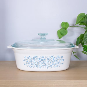 May include: White casserole dish with a clear glass lid. The dish has a floral pattern in light blue. The lid has a knob handle. The dish is on a wooden surface.