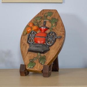 May include: A wooden, oval-shaped wall plaque featuring a red coffee grinder with black wheels and a black base. The plaque has a brown background with orange fruit and green leaves. The plaque is supported by a wooden stand.