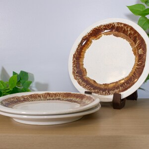 May include: A set of vintage ceramic plates with a cream-colored center and a wide, brown, textured border. Two plates are stacked, and one is displayed on a wooden stand. The plates are on a wooden surface with green foliage in the background.