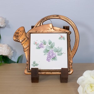 May include: A decorative, vintage-style tile in a gold-colored metal frame shaped like a watering can. The tile features a grape cluster design in purple, green, and white. The frame is supported by two wooden blocks, and the background is a light blue wall.