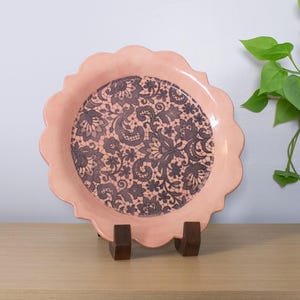 May include: A decorative, scalloped-edge plate in a soft peach color. The center features a dark gray lace pattern. The plate is displayed on a wooden stand, showcasing its intricate design. A unique piece for home decor.