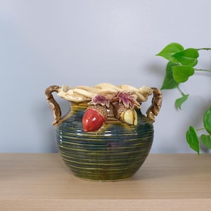 May include: A ceramic vase with a ribbed, dark green and blue body. The rim and handles are tan, with decorative acorns and leaves in red, tan, and purple. The vase is on a light brown surface.
