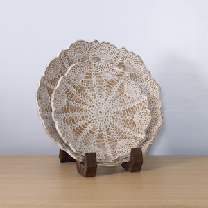 May include: Two decorative plates with a white lace design, displayed on a wooden stand. The plates have a beige background and a scalloped edge. The stand is made of dark brown wood.