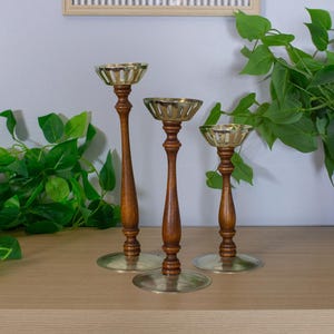 May include: Three vintage candle holders with wooden stems and brass-colored tops and bases. The candle holders are of varying heights and are arranged on a light-colored wooden surface. Green plants are in the background.