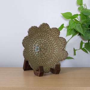 May include: A decorative, scalloped-edge bowl with a textured, speckled surface in shades of brown and green. The bowl is displayed on a dark wooden stand. A green plant is in the background.