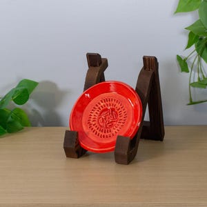 May include: A bright red ceramic dish with a detailed embossed design, displayed on a dark brown wooden stand. The dish is round with a textured surface. The stand has a rustic, handcrafted appearance.