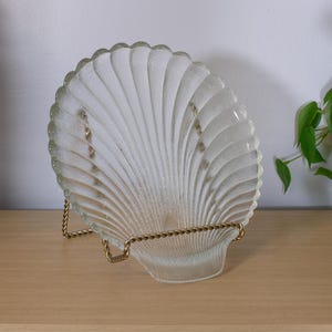 May include: A clear glass dish shaped like a scallop shell with a textured, ribbed surface. The dish is held upright by a gold-toned metal wire display stand, showcasing the translucent material and the distinct fan-like ridges of the shell design.