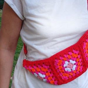 Può includere: Un marsupio rosso all'uncinetto con cinturino, caratterizzato da un colorato design a quadretti granny. I quadrati incorporano motivi floreali rosa, arancioni e bianchi. La borsa è indossata a tracolla.