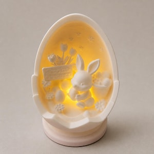 Könnte beinhalten: Eine weiße, eiförmige Porzellanleuchte mit einem Relief aus einem Hasen, Blumen und kleinen Sternen im Inneren. Das Innere ist in ein warmes gelbes Licht getaucht, das die detaillierte Hasenfigur und den Schriftzug Frohe Ostern hervorhebt.