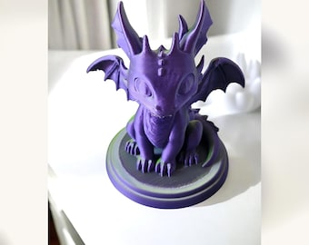 May include: A small, purple, 3D-printed dragon figurine with large eyes, pointed ears, and wings, sitting on a circular base. The creature has a textured surface with visible layer lines, a small tail, and a detailed head with small horns.