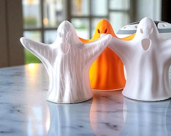 May include: Three ghost-shaped figurines standing in a row. The left figure is pearlescent white, the center figure is bright orange, and the right figure is matte white. Each ghost features cut-out eyes and an open mouth, with arms outstretched.