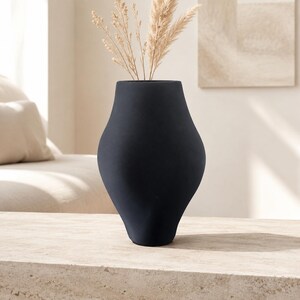 May include: A matte black vase with a curved, organic silhouette and a wide, rounded body that tapers toward the base and the opening. The vase contains several tall, dried beige pampas grass stems extending from the top opening.