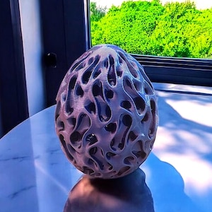 Könnte beinhalten: Ein 3D-gedrucktes, eiförmiges Objekt mit einer komplexen, hohlen, organischen Gitterstruktur. Die Oberfläche hat ein metallisches, hellviolettes Finish mit sichtbaren horizontalen Schichtlinien im gesamten Design.