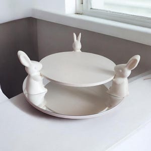 Könnte beinhalten: Ein weißer, zweistöckiger Servierständer aus Keramik mit Hasenfiguren. Die untere Ebene ist eine flache Schale, und die obere Ebene ist ein kleinerer, runder Teller. Eine kleine Hasenfigur befindet sich oben.