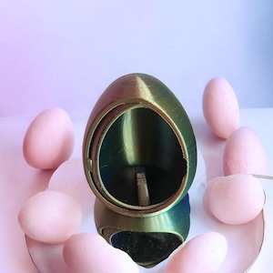 Può includere: Un oggetto metallico verde a forma di uovo con un'apertura circolare, circondato da diversi oggetti rosa a forma di uovo. Gli oggetti sono disposti su una superficie bianca e rosa, creando una composizione visivamente accattivante.