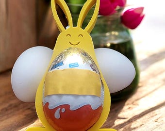 May include: A bright yellow 3D-printed rabbit-shaped holder featuring a smiling face cutout. The holder contains a foil-wrapped chocolate egg in the center and two white oval-shaped objects positioned on either side to resemble bunny arms.