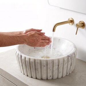 Puede incluir: Un lavabo de sobreponer de mármol blanco con diseño exterior acanalado, combinado con un grifo de pared en oro cepillado y un mando monomando a juego. El lavabo presenta vetas grises naturales y está colocado sobre una encimera color hormigón.
