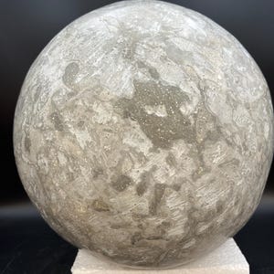 May include: A large, spherical decorative object with a textured, mottled surface in shades of gray and white. The sphere is resting on a white foam base, set against a dark background. The overall appearance suggests a celestial or geological theme.