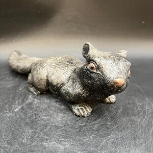 Puede incluir: Una detallada figura de una ardilla en posición sentada. La escultura es principalmente gris con sombras más oscuras y una superficie texturizada. La ardilla tiene una cola esponjosa, orejas pequeñas y una cara detallada con ojos marrones y una nariz rosada. La figura está sobre una superficie oscura.
