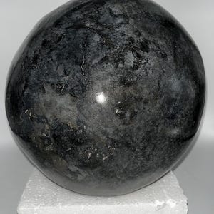 May include: A dark, rounded stone sculpture with a mottled, textured surface. The stone is primarily shades of gray and black, with subtle variations in color and pattern. The sculpture is displayed on a white foam base.
