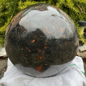May include: A large, spherical, dark-colored decorative object with a glossy finish. The surface has a mottled appearance with reddish-orange accents. The object is resting on a white towel, with green foliage in the background. The sphere appears to be made of stone.