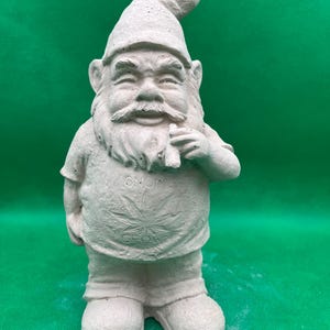 May include: A concrete garden gnome statue. The gnome wears a pointed hat, a t-shirt with a cannabis leaf design, and holds a rolled item. The statue is light gray and stands on a green surface.