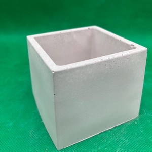 Puede incluir: Una maceta cuadrada de hormigón gris claro con un exterior liso. La maceta tiene un diseño simple y moderno y es adecuada para plantas pequeñas o suculentas. El interior es de un tono gris más claro.
