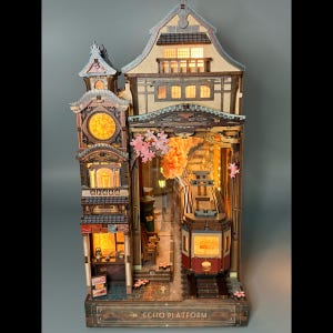 May include: A wooden book nook diorama titled Echo Platform featuring a multi-story building with a clock tower, illuminated windows, and a streetcar on tracks. The structure includes pink cherry blossom accents and warm interior lighting.