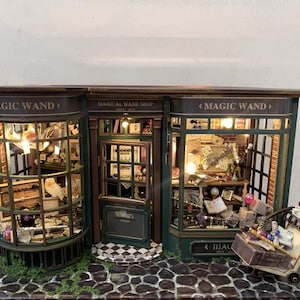 Könnte beinhalten: Miniaturmodell eines Zauberstabladens mit detaillierten Innen- und Außendetails. Die Fassade des Ladens ist grün und braun, mit den Worten "MAGIC WAND" und "MAGICAL WAND SHOP". Ein kleines Schild zeigt "MAGIC WAND". Ein Wagen ist mit Kisten gefüllt.