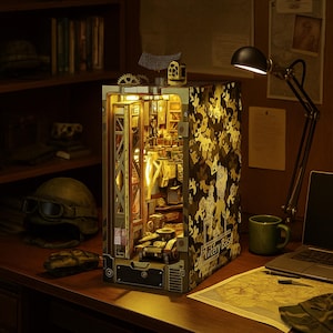 May include: A detailed, illuminated diorama of a military base, featuring a camouflage exterior and intricate interior details. The model includes miniature vehicles and structures, with the text "Phitery Base" visible on the side. The scene is set on a wooden desk with a lamp and map.