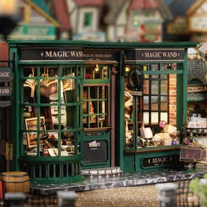 May include: A miniature green and black "Magic Wand" shop with detailed architectural features. The shop has large display windows filled with books and magical items. The sign reads "Magic Wand Shop". A small barrel and a luggage cart are visible.