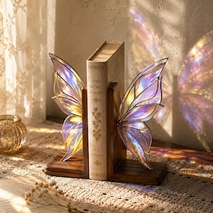 Può includere: Una coppia di fermalibri in legno scuro con ali di fata in vetro colorato iridescente con bordi metallici. Le ali presentano una finitura scintillante e multicolore che riflette la luce, posizionate ai lati di un libro intitolato The Secret Garden.
