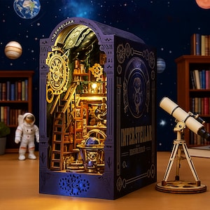 May include: A detailed wooden book nook with an interstellar theme. The interior features a miniature room with a ladder, globe, and intricate details, illuminated by warm lighting. The exterior is dark blue with gold accents and the word "INTERSTELLAR". A small telescope sits nearby.