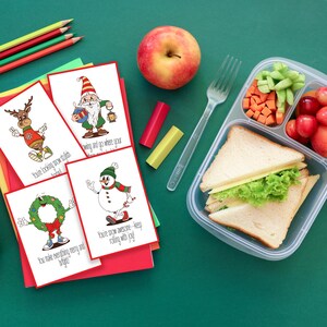 May include: A festive lunch scene with Christmas-themed cards, a red apple, and a packed lunch. The cards feature cartoon characters with holiday greetings. The lunchbox contains a sandwich, vegetables, and fruit. Colored pencils and crayons are also present.
