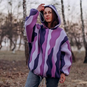 Pode incluir: Casaco com capuz e zíper com um padrão ondulado em tons de roxo e lavanda. O casaco tem um cordão no capuz e na cintura. A pessoa está usando jeans cinza. O fundo é uma cena externa desfocada com árvores.