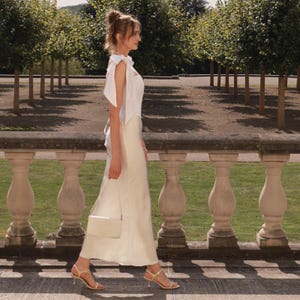 K&ouml;nnte beinhalten: Ein wei&szlig;es &auml;rmelloses Kleid mit einem Oberteil mit Bindetr&auml;gern und einem bodenlangen Satinrock. Das Ensemble wird mit einer kleinen wei&szlig;en rechteckigen Umh&auml;ngetasche und hellen Riemchensandalen mit hohem Absatz kombiniert.