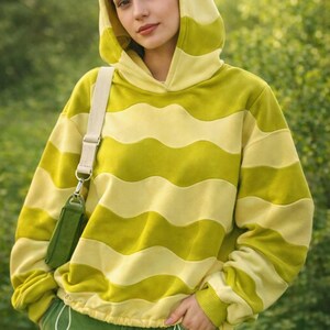 May include: A hooded sweatshirt with horizontal wavy stripes in shades of yellow and green. The drawstring waist and cuffs match the green pants. A small green purse hangs from a cream-colored strap.