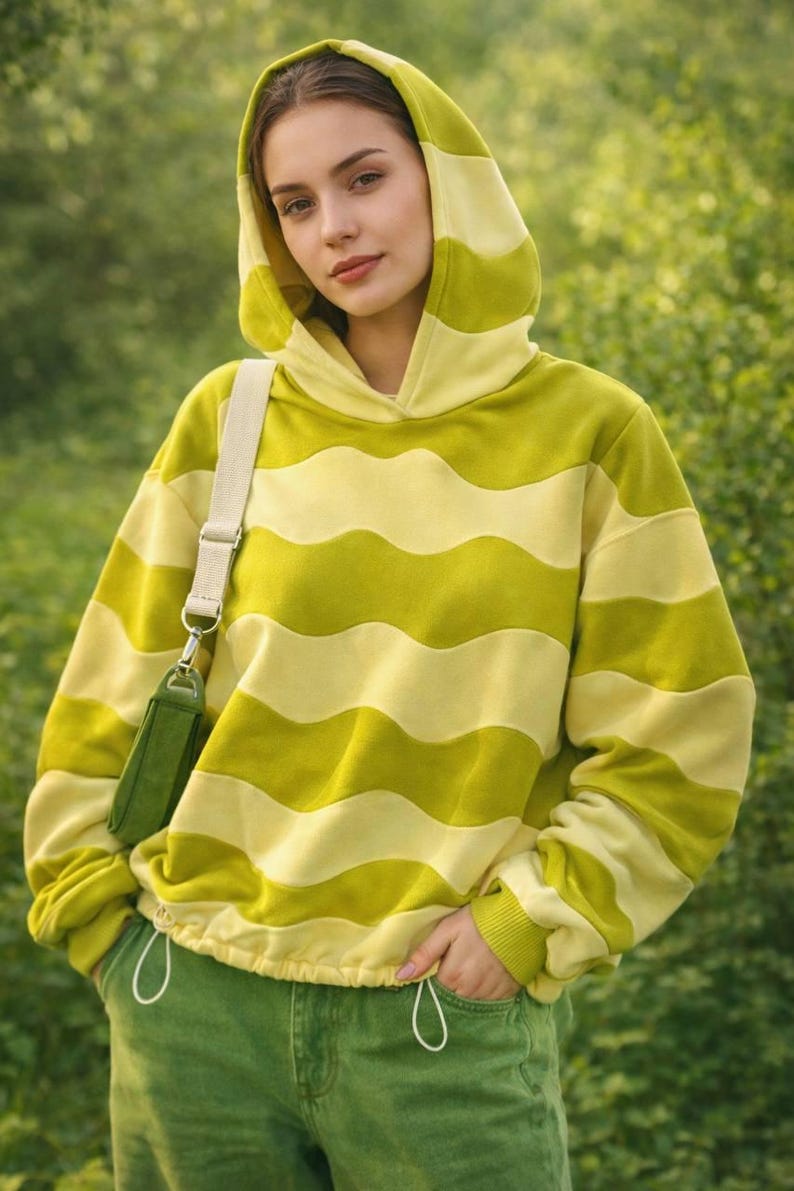 May include: A hooded sweatshirt with horizontal wavy stripes in shades of yellow and green. The drawstring waist and cuffs match the green pants. A small green purse hangs from a cream-colored strap.
