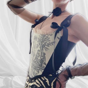 Può includere: Un corsetto nero e crema con una stampa toile de Jouy raffigurante un albero e delle figure. Il corsetto ha spalline nere con fiocchi, un choker nero con una rosa ed è abbinato a una gonna con stampa zebrata.