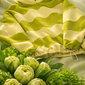 May include: A green and cream-colored hooded sweatshirt with a wavy stripe design. The sweatshirt is lying on a green surface next to a bouquet of green flowers and leafy greens.