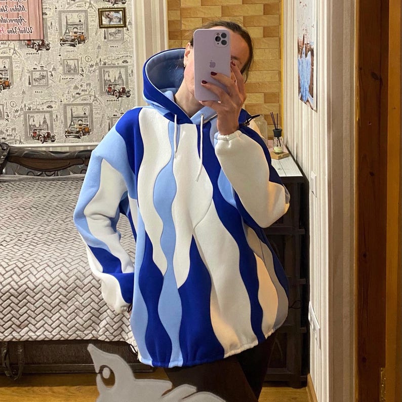 May include: A blue, white, and light blue hoodie with a unique wavy design. The hoodie has a drawstring hood and white cuffs. The design is a bold, abstract pattern. The hoodie is made of a soft material.